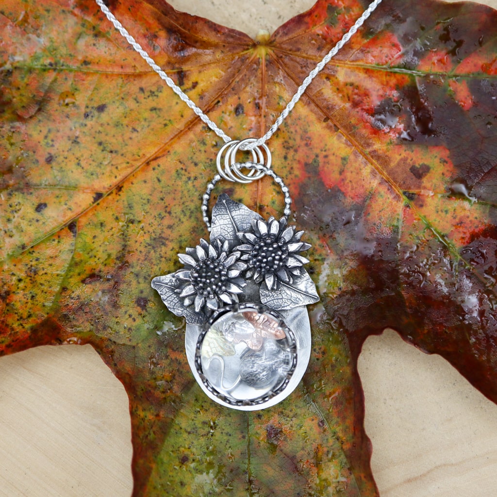 A sterling silver mouse is tucked under a glass dome and lives amongst some silver, copper, and brass fall leaves. You can shake the pendant and the mouse and leaves change their position. Above them are two silver sunflowers and their leaves. The pendant is about 2.25 inches tall and shown on a green, orange, red, and black fall leaf.