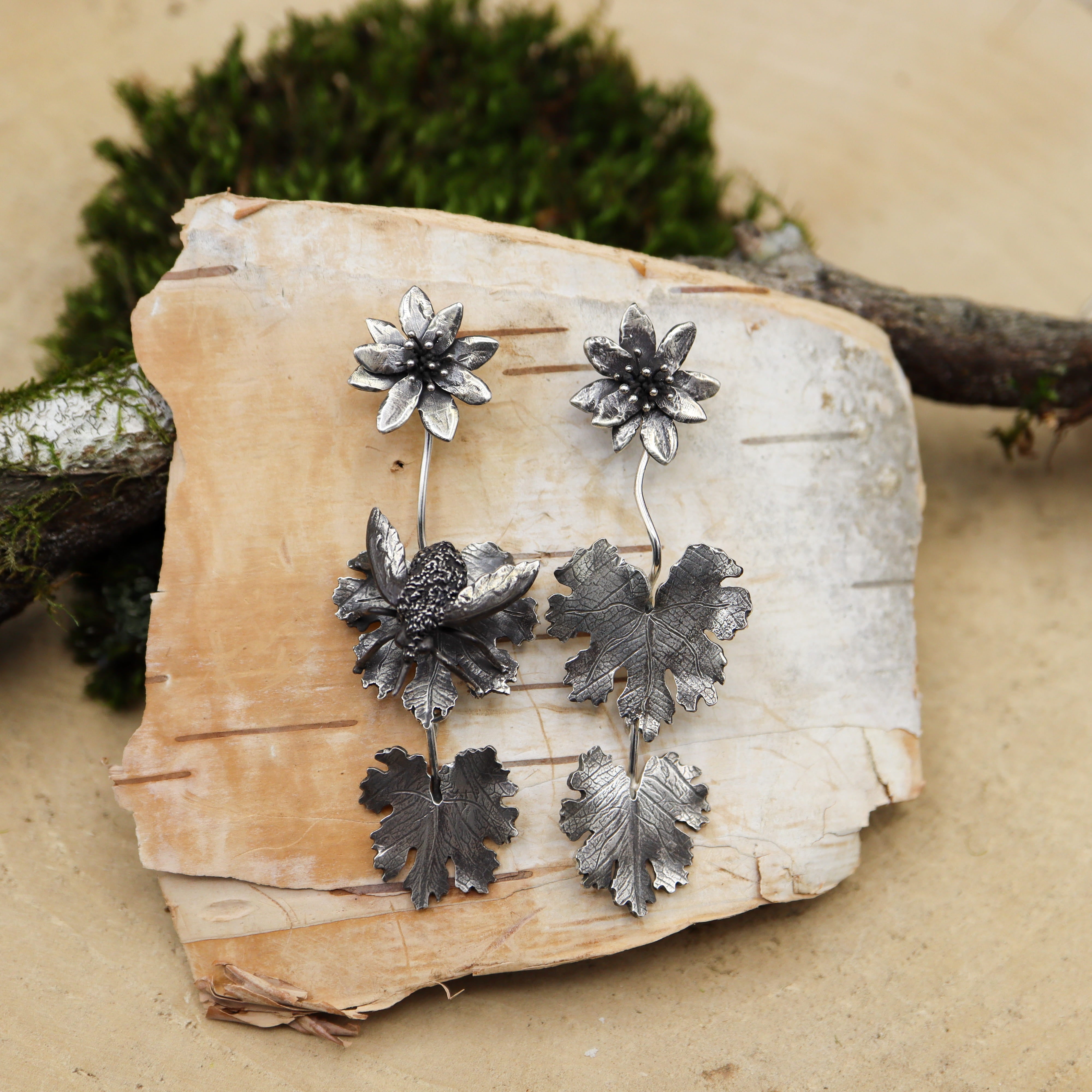 A pair of sterling silver earrings featuring bloodroot flowers. One of the earrings has a hand carved bee fly on the leaf. They are shown on a piece of white birch bark.