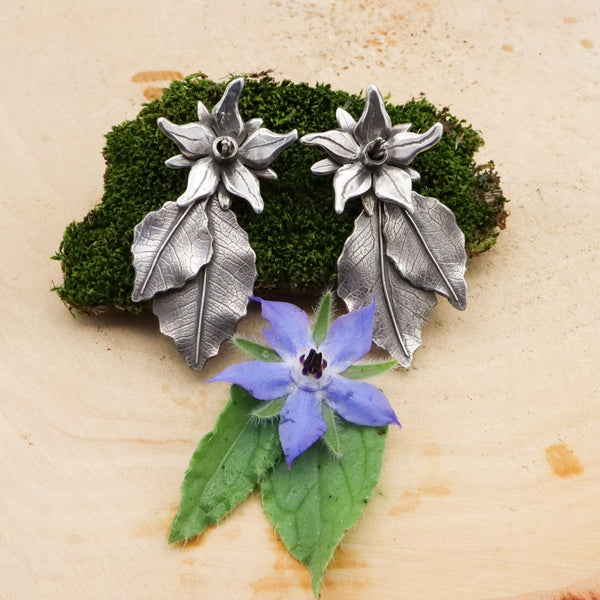 Sterling silver borage flower and leaf earrings. The two dangle earrings are shown on a piece of dark green moss with a real borage flower below them. 