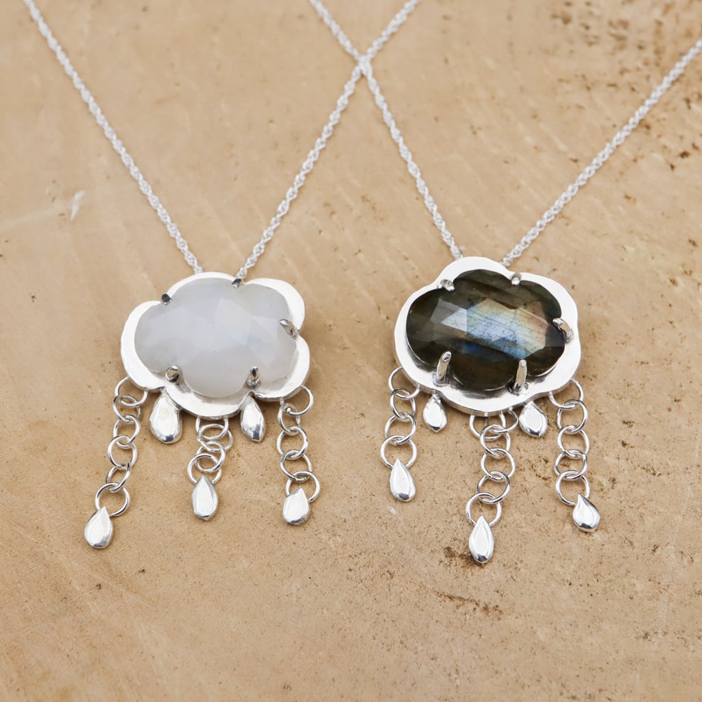 A happy moonstone raincloud necklace next to a sad labradorite raincloud necklace. Both necklaces have sterling silver dangles below them with tiny raindrops attached. They are both shown on a piece of light tan wood. 