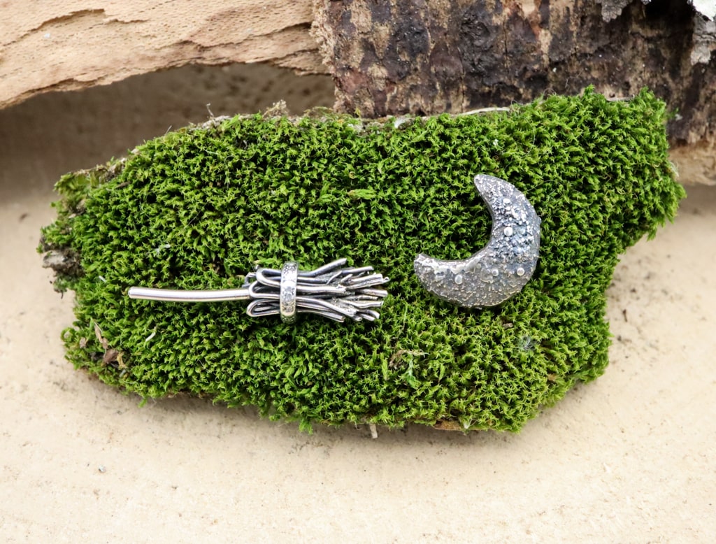 A close up of the witches broom and crescent moon stud earrings. They are shown on a piece of dark green moss. 