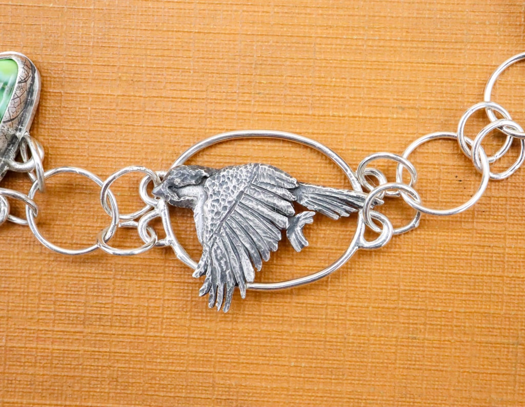 A close up view of the hand carved sterling silver chickadee bird on the right side of the bracelet. It is shown on a bright orange vintage book. 