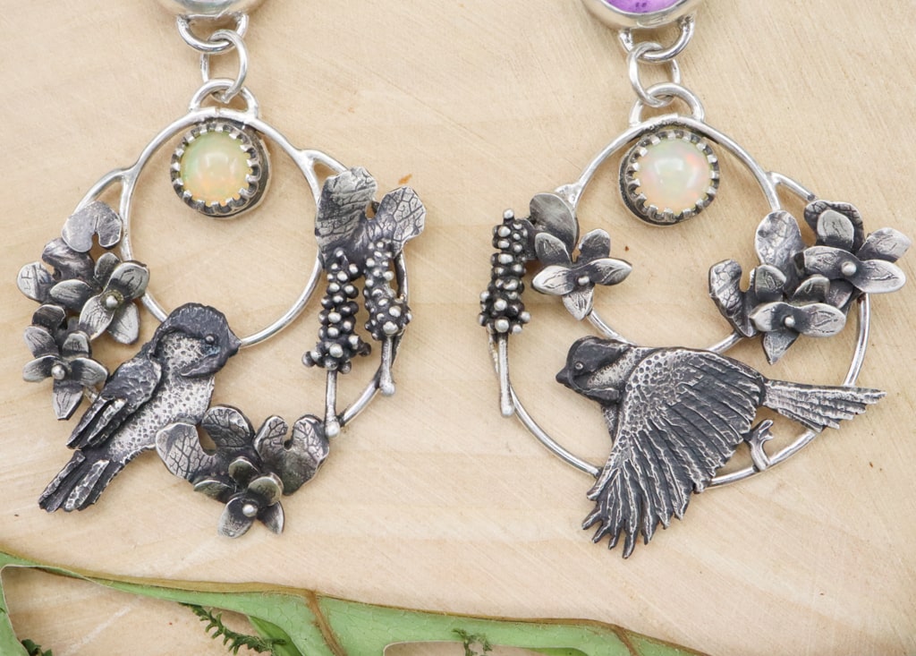 A closer view of the handmade sterling silver chickadee earrings featuring violet flowers, leaves, and hyacinth flowers. They are shown on a piece of light tan wood. 