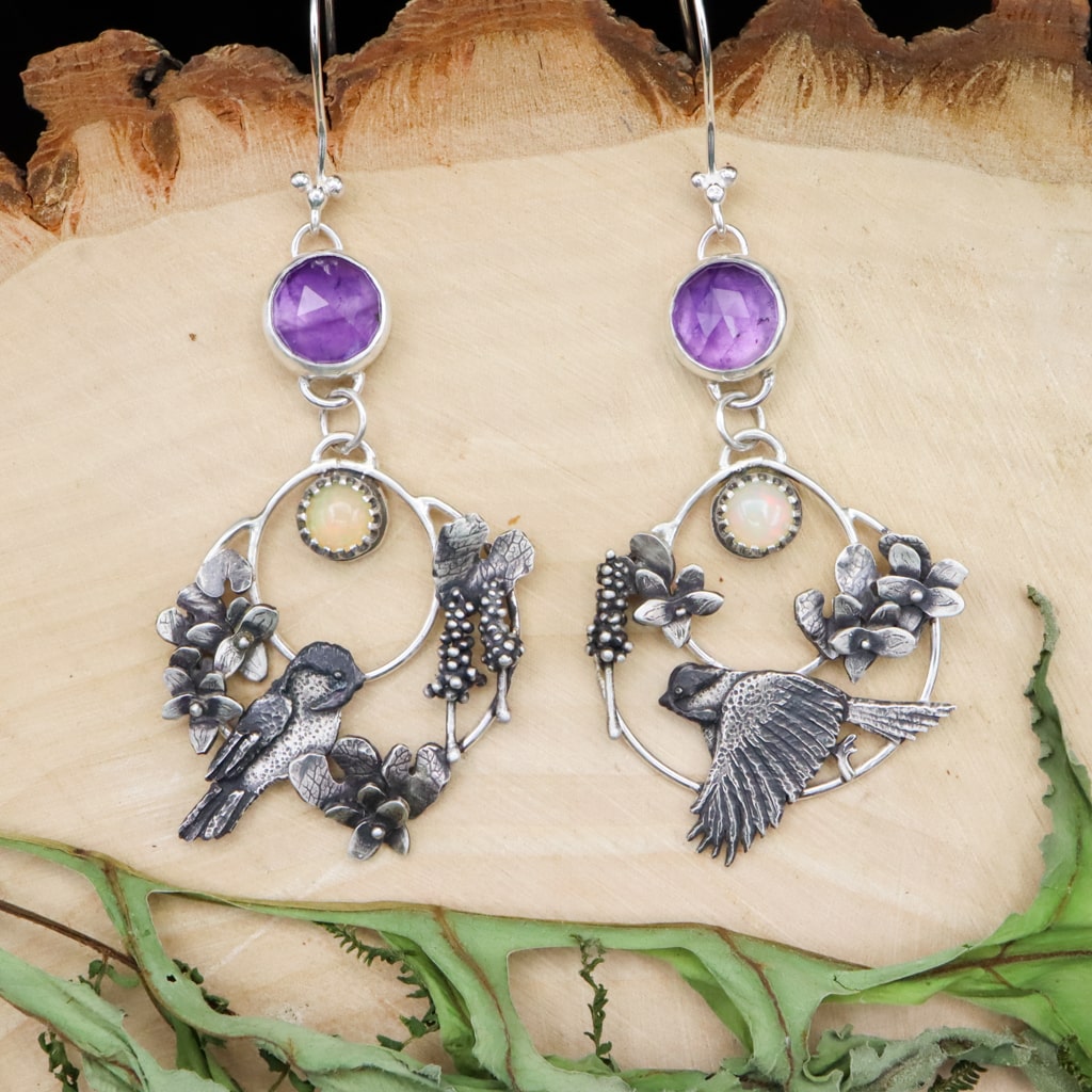 Sterling silver handmade chickadee bird earrings are shaped into hoops. They have an opal stone and amethyst stone at the top. The chickadees are surrounded by tiny violet flowers, leaves, and hyacinth flowers. The earrings are shown on top of a piece of light tan wood. 