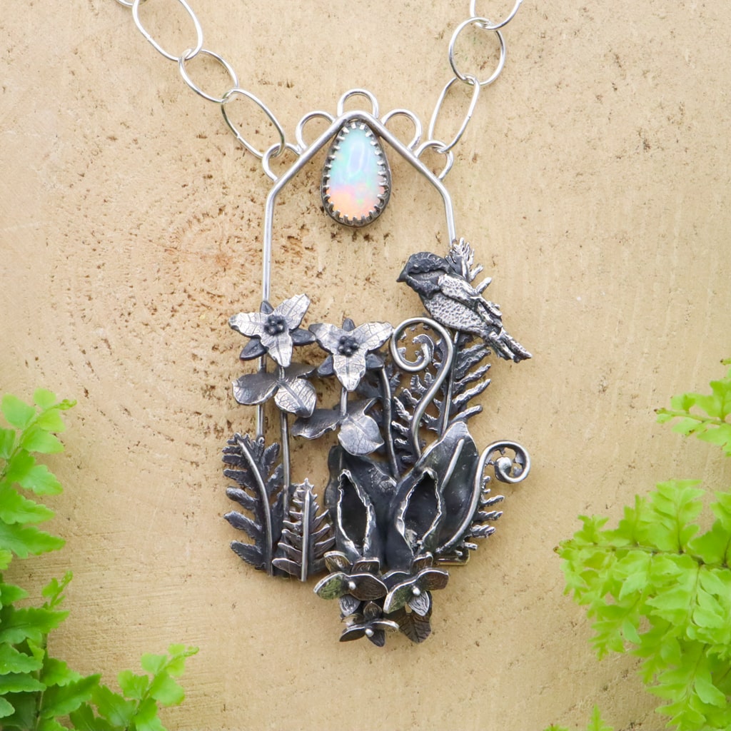 A handmade sterling silver necklace that is sort of shaped like a window. Within the window frame are lots of ferns, some violet flowers and heart shaped leaves, 2 skunk cabbage flowers, and two trillium flowers. At the top of one of the ferns is a little chickadee bird perched. At the top of the necklace is a pear shaped rainbow colored opal..
