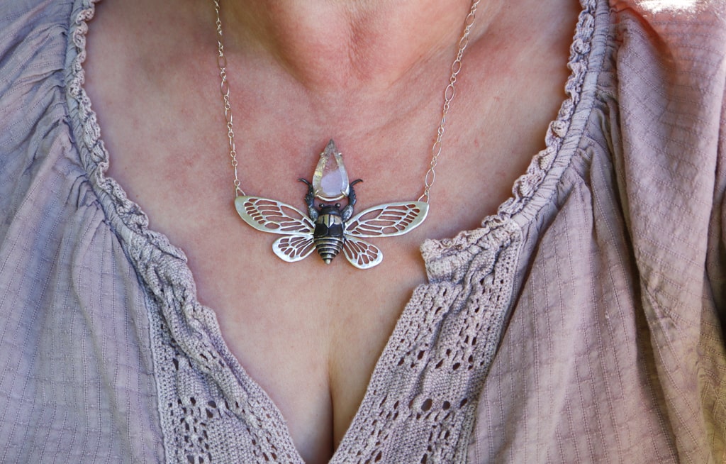 A sterling silver cicada necklace is shown being worn by a woman in a light purple blouse. 