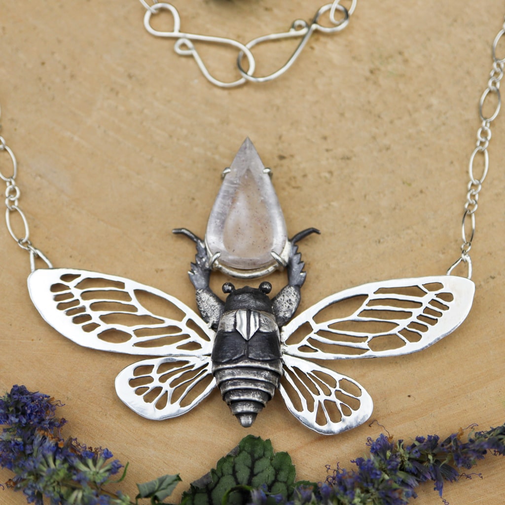 A handmade sterling silver woman's necklace featuring a cicada. he cicada has a teardrop shaped covellite in quartz gemstone above him. It is shown on a light tan piece of wood with some purple flowers below it. 
