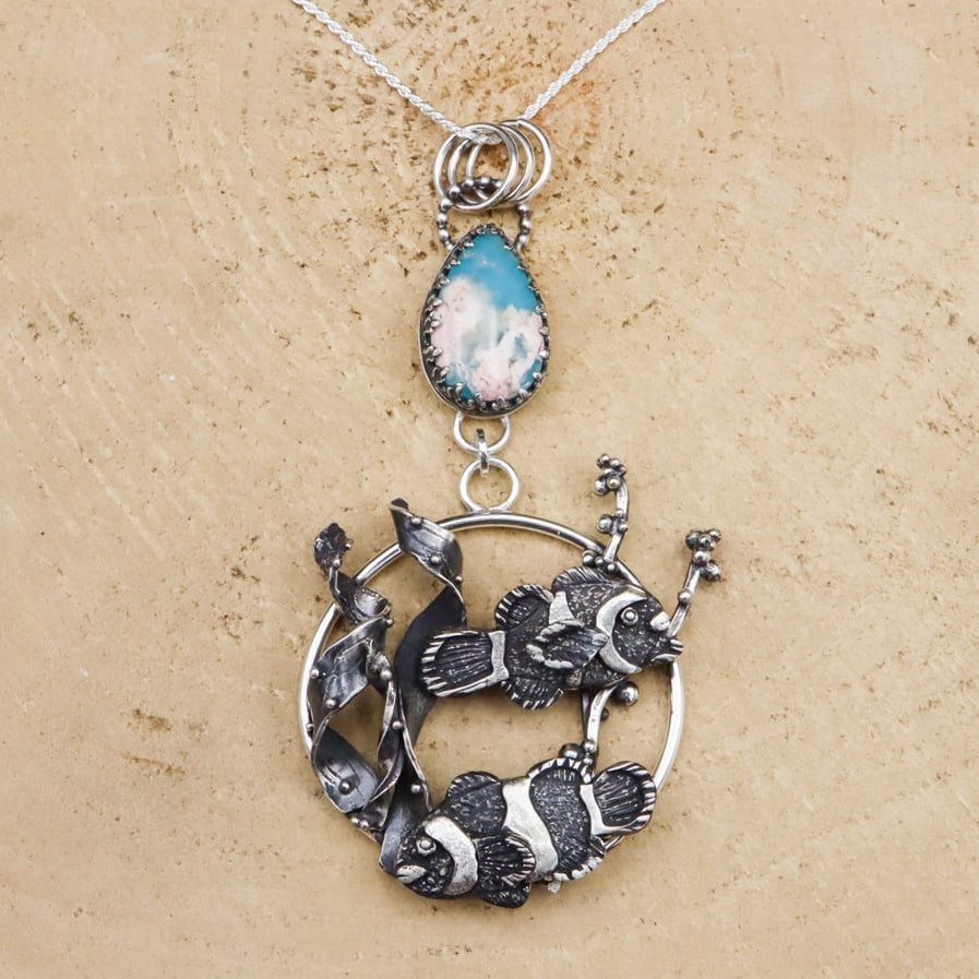 A sterling silver necklace featuring a pair of clownfish with white stripes, accompanied by a turquoise and agate doublet stone, on a wooden surface.