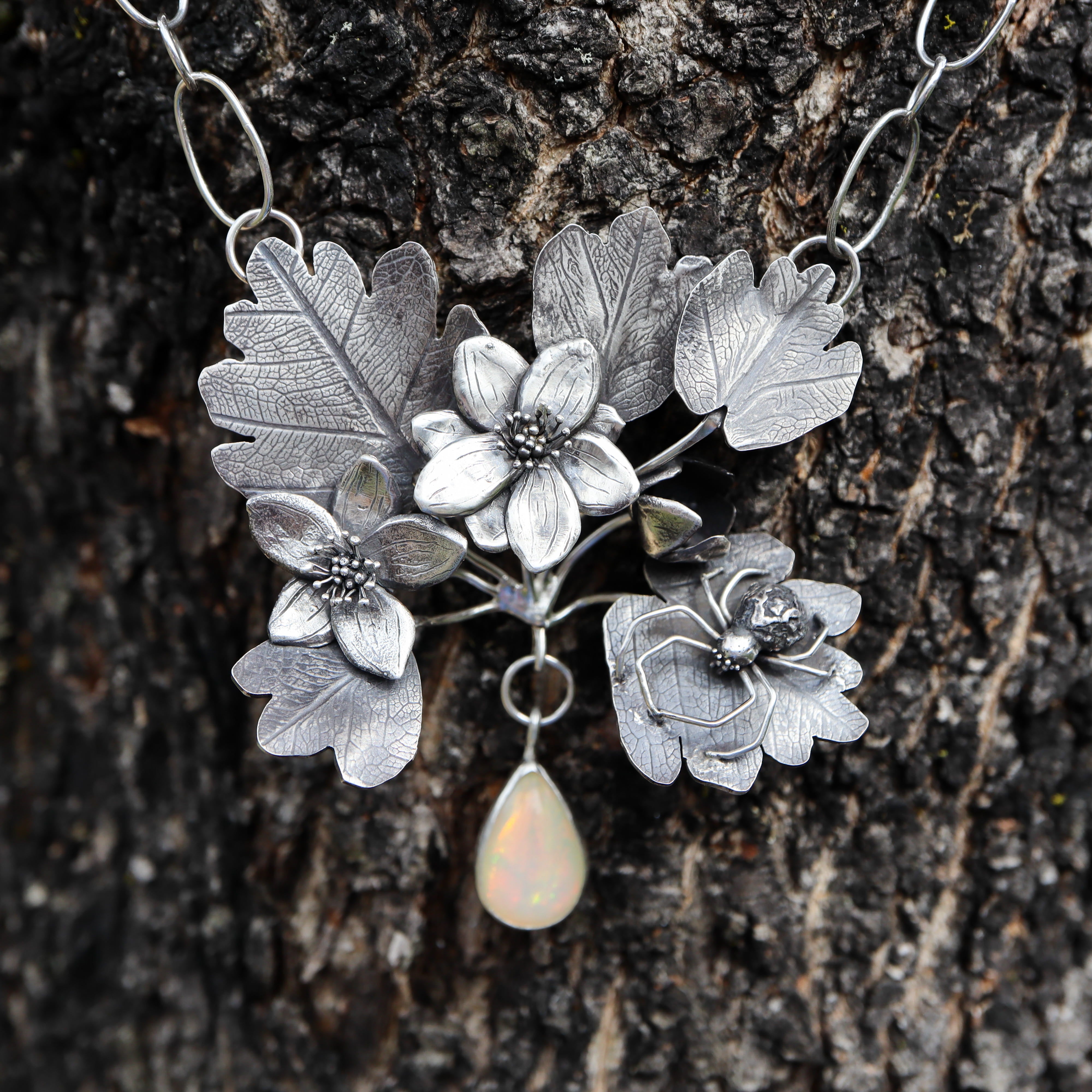 A handmade sterling silver rue anemone necklace with a little crab spider sitting on one of the leaves. The statement necklace is shown on the dark bark of a tree. 
