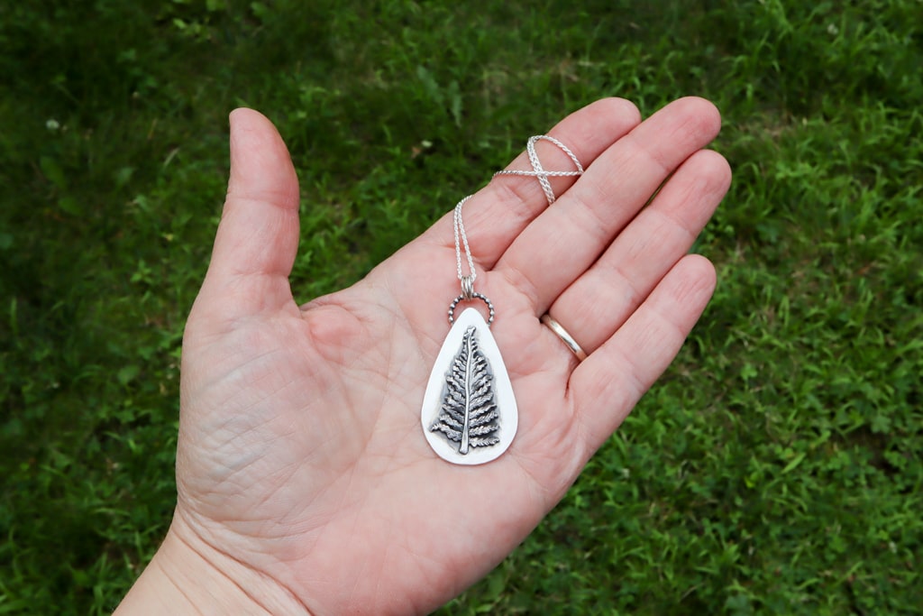 A hand is shown holding the handmade sterling silver fern necklace for size reference. The pendant is about 2 inches tall. 