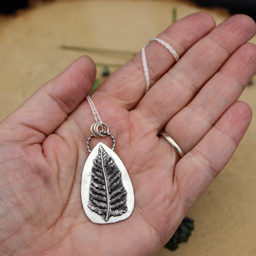 A hand is shown holding the handmade sterling silver fern necklace. 