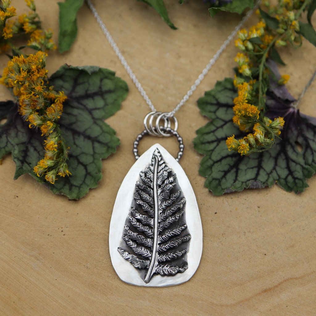 A handmade sterling silver fern necklace. It is about 1.5 inches tall and hand carved so each one is one of a kind. It is shown on a light tan piece of wood with yellow goldenrod flowers around it. 