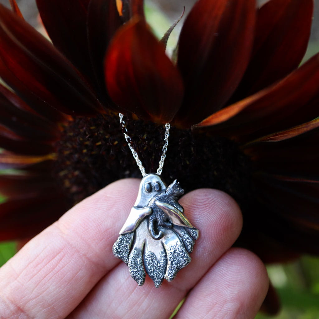 A silver pendant necklace featuring a ghost holding a small cat, held in a person's hand in front of a dark sunflower.