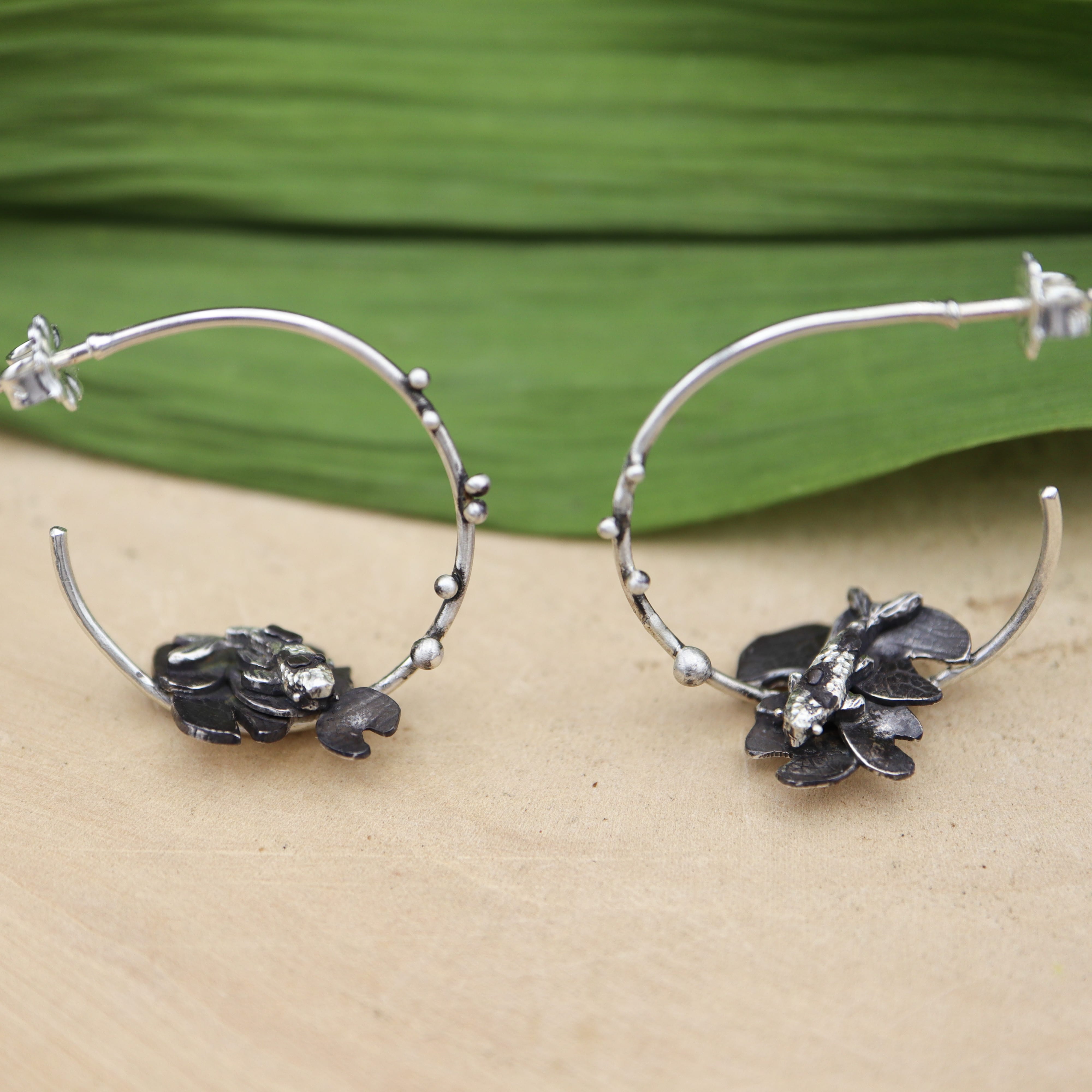 Silver handmade goldfish hoop earrings. They are shown on a piece of light tan wood with a green leaf behind them. 