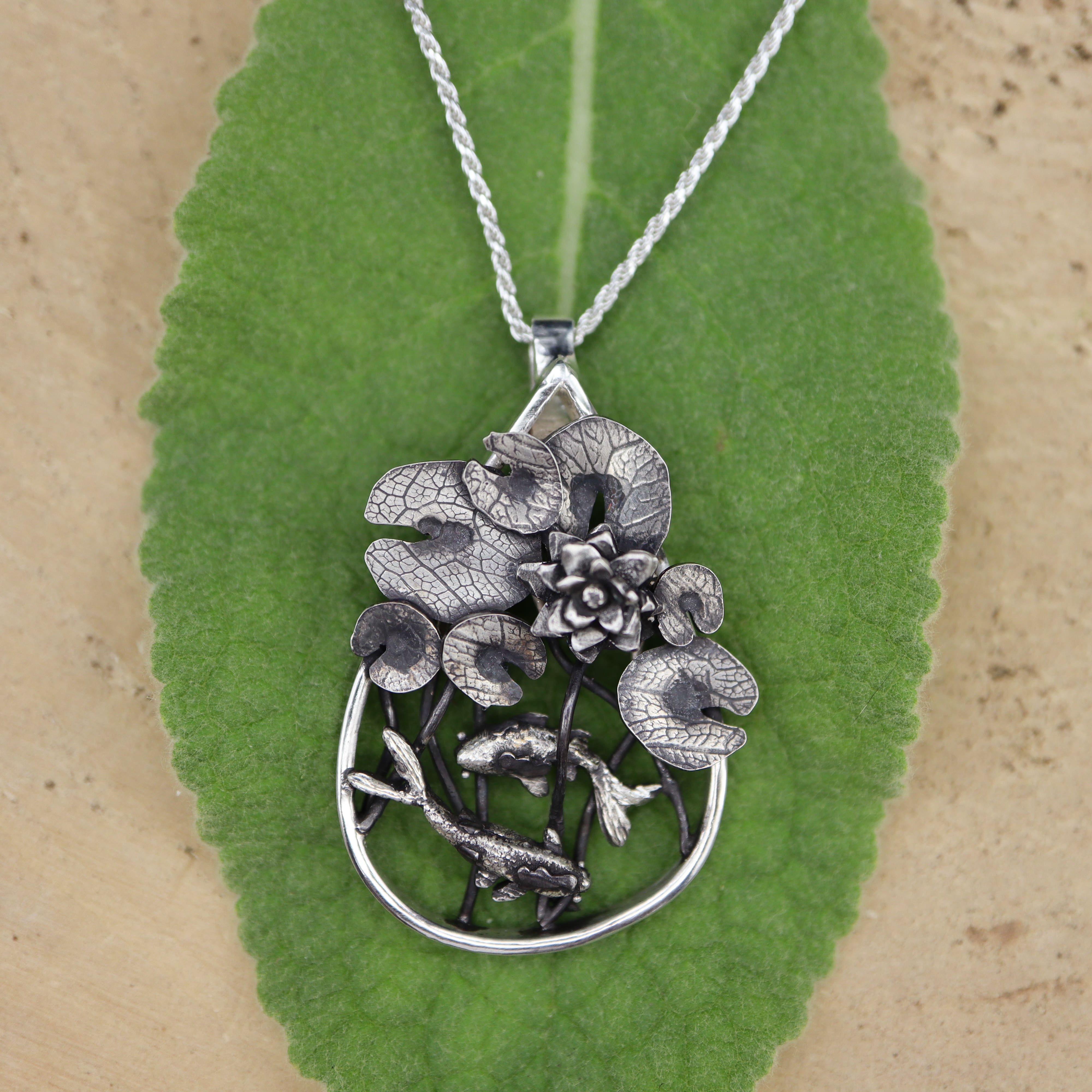 Silver lily pad and goldfish pendant necklace on a green leaf with a brown background