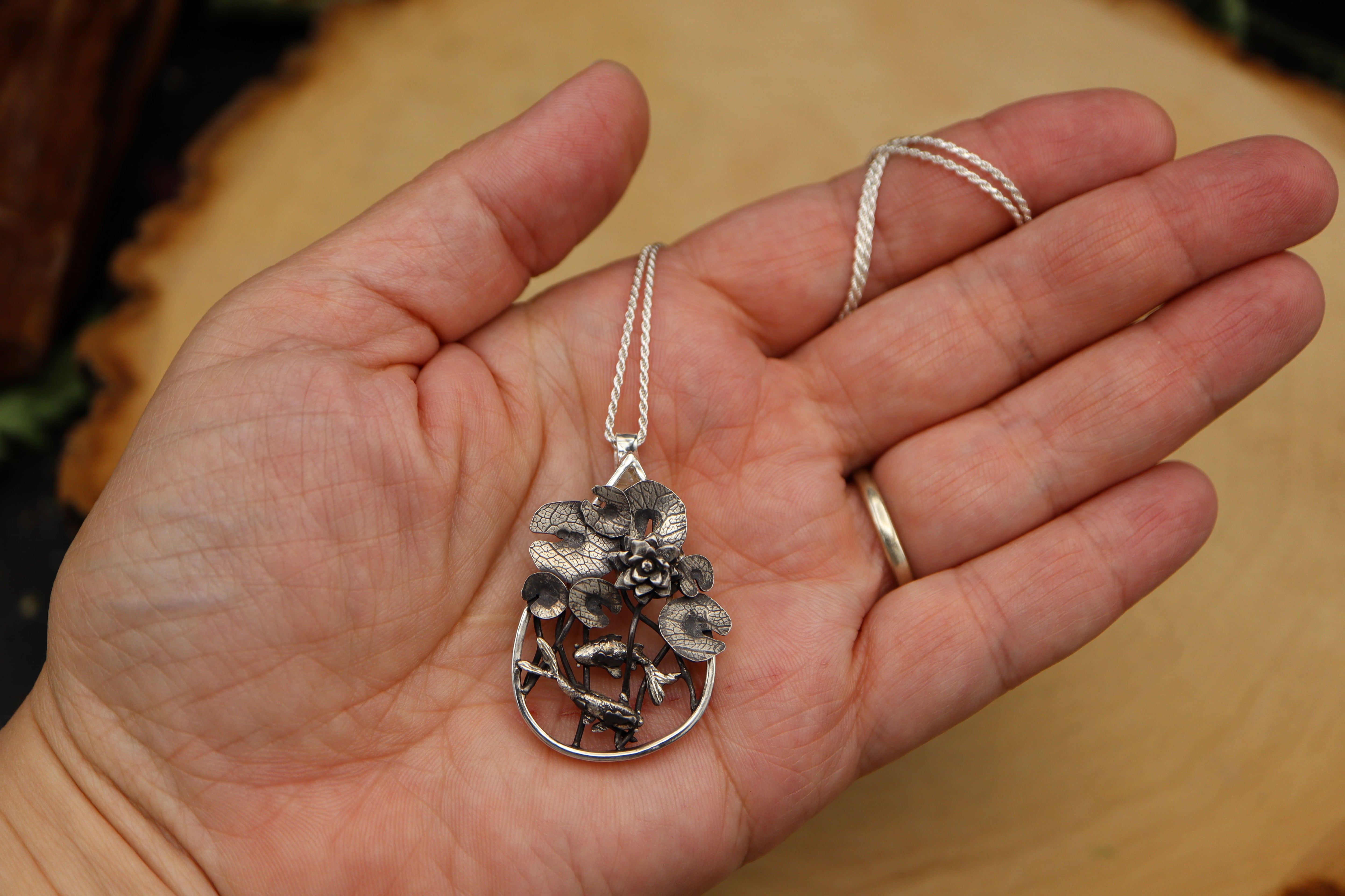 Hand holding a silver pendant with a natural design on a wooden surface