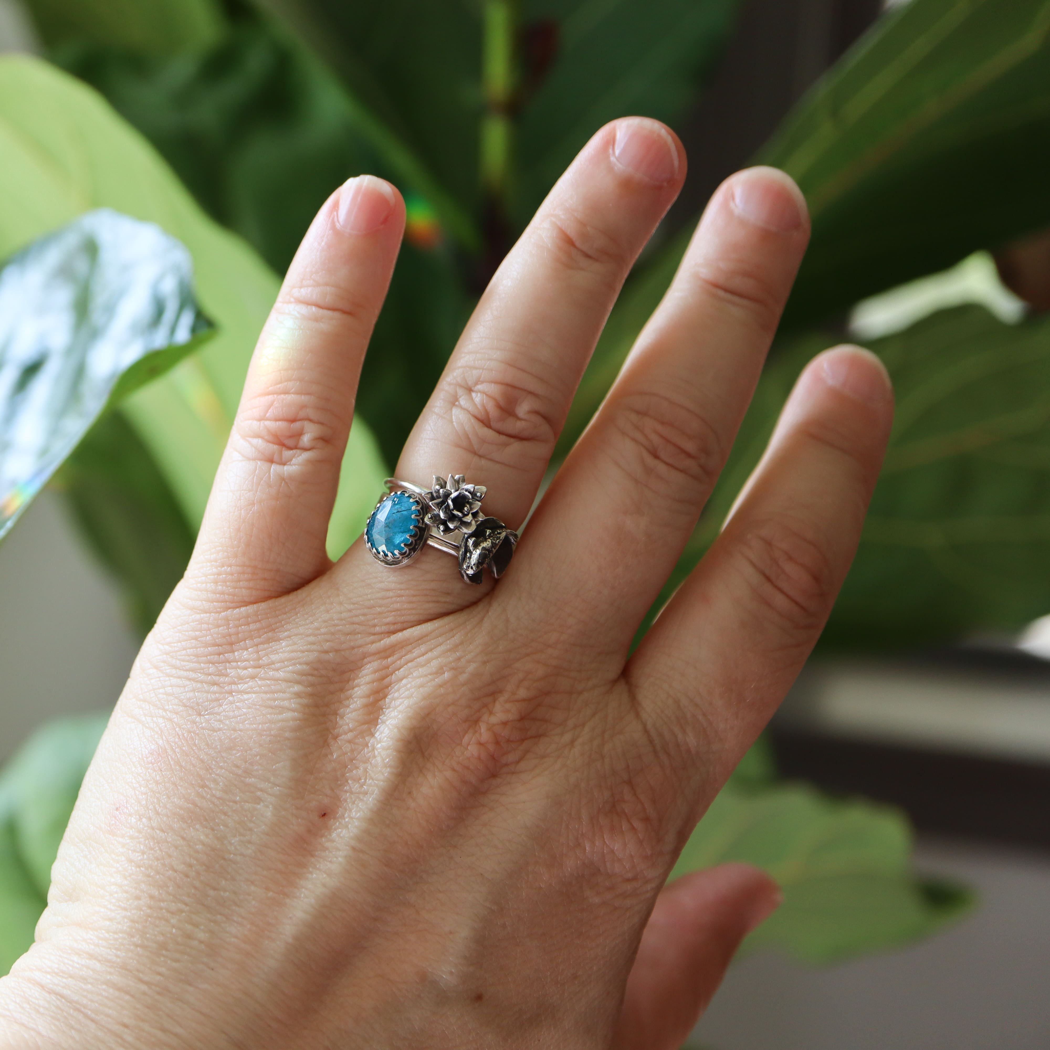 Hand wearing a ring with a blue apatite gemstone, water lily flower, and goldfish band against a green leafy background