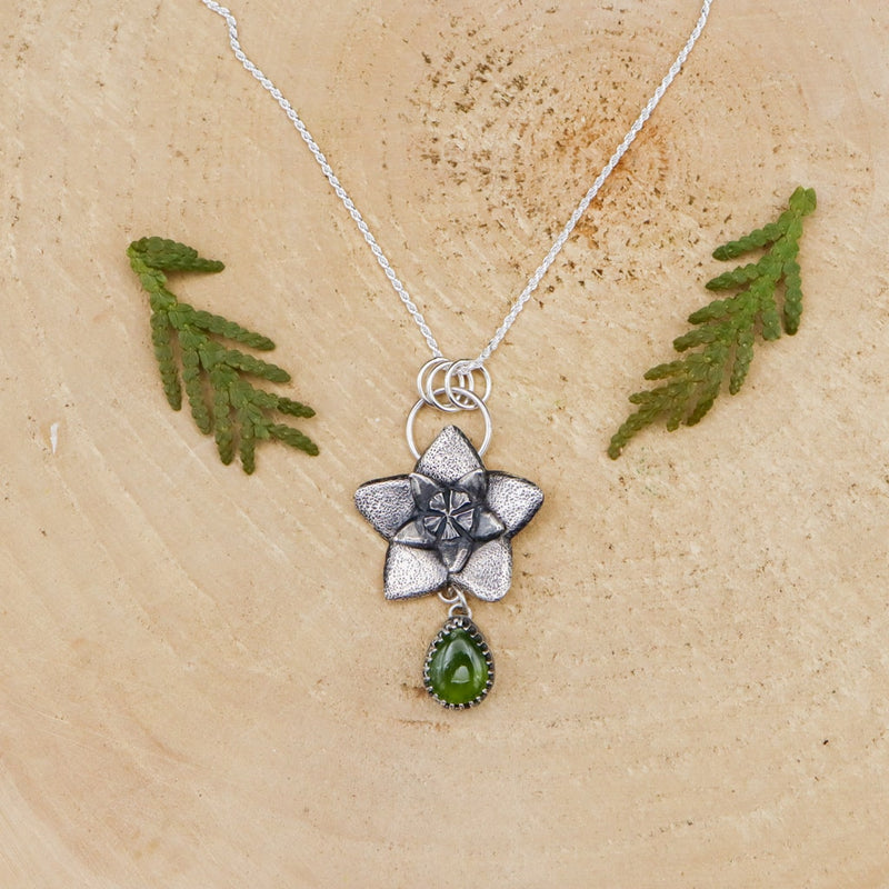 Hoya flower bud necklace is shown on a light tan piece of wood. The flower is about 1 inch tall and below it is a dark green pear shaped vesuvianite stone.