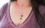 The necklace is being shown worn by a woman with dark brown hair that is long and a dark purple shirt. 