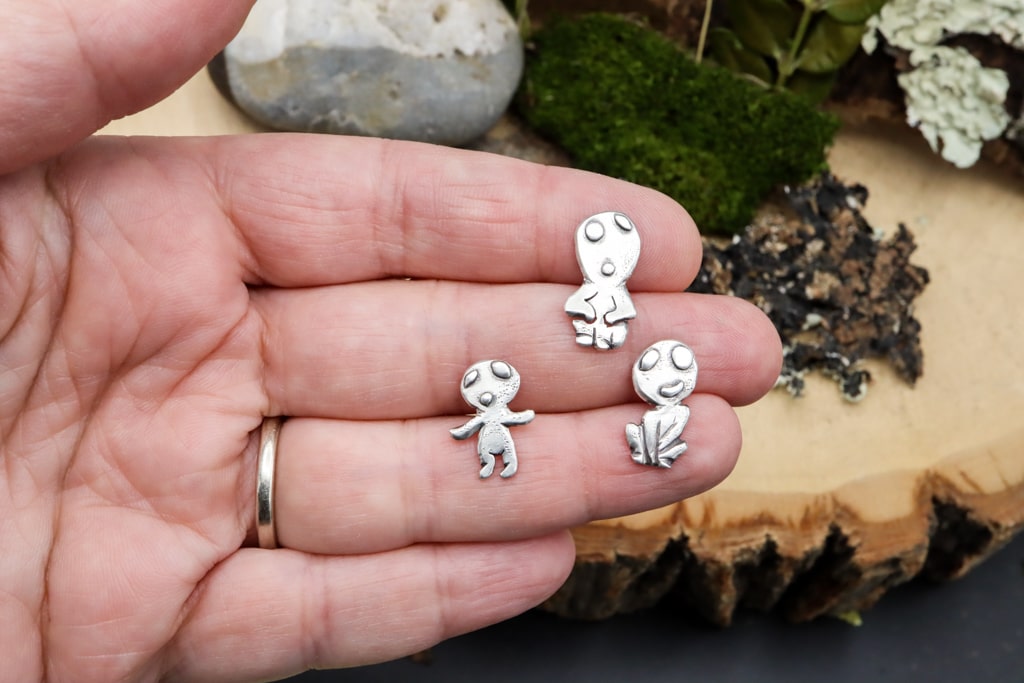 A hand is shown holding the sterling silver kodama for size reference. There are three of them. One is sitting with his hands on his knees, one is standing with arms outstretched, and the third is sitting cross legged. 
