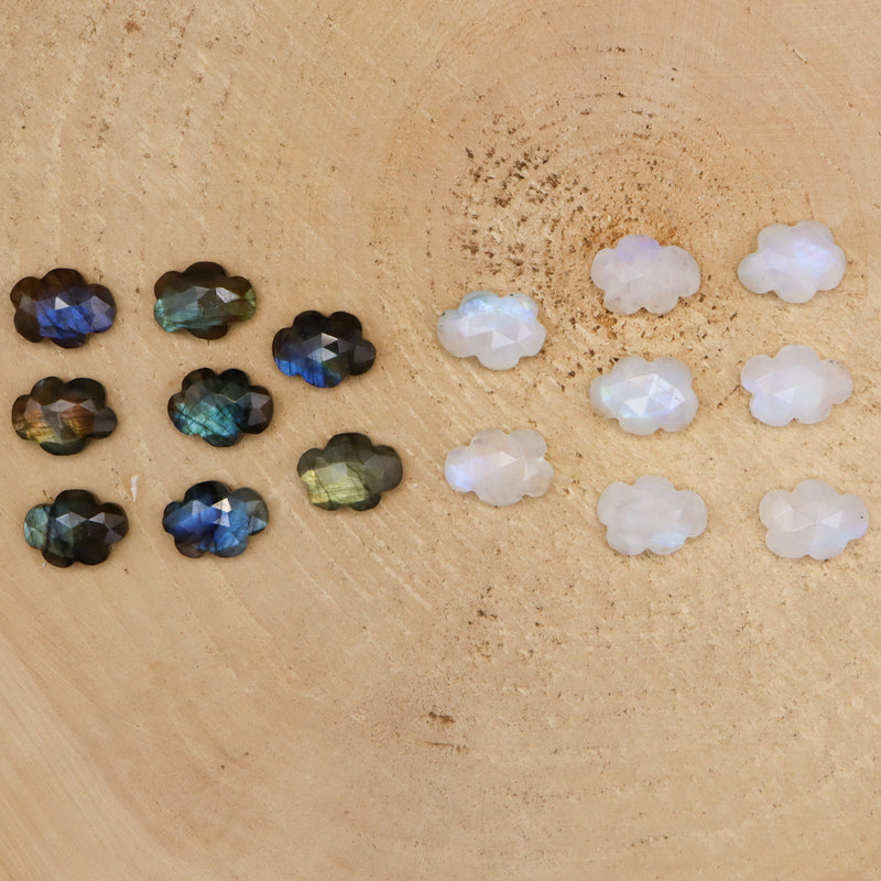 Eight labradorite stones that are shaped like little clouds are next to eight cloud shaped rainbow moonstones. They are all shown on a piece of light tan wood. 
