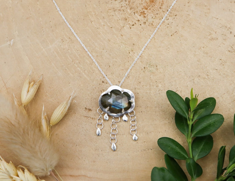 A labradorite raincloud necklace is shown on a piece of light tan wood. The pendant has a stone that is cut in the shape of a cloud. Below the cloud are silver loops connecting in a chain and tiny silver raindrops at the bottom of them. 