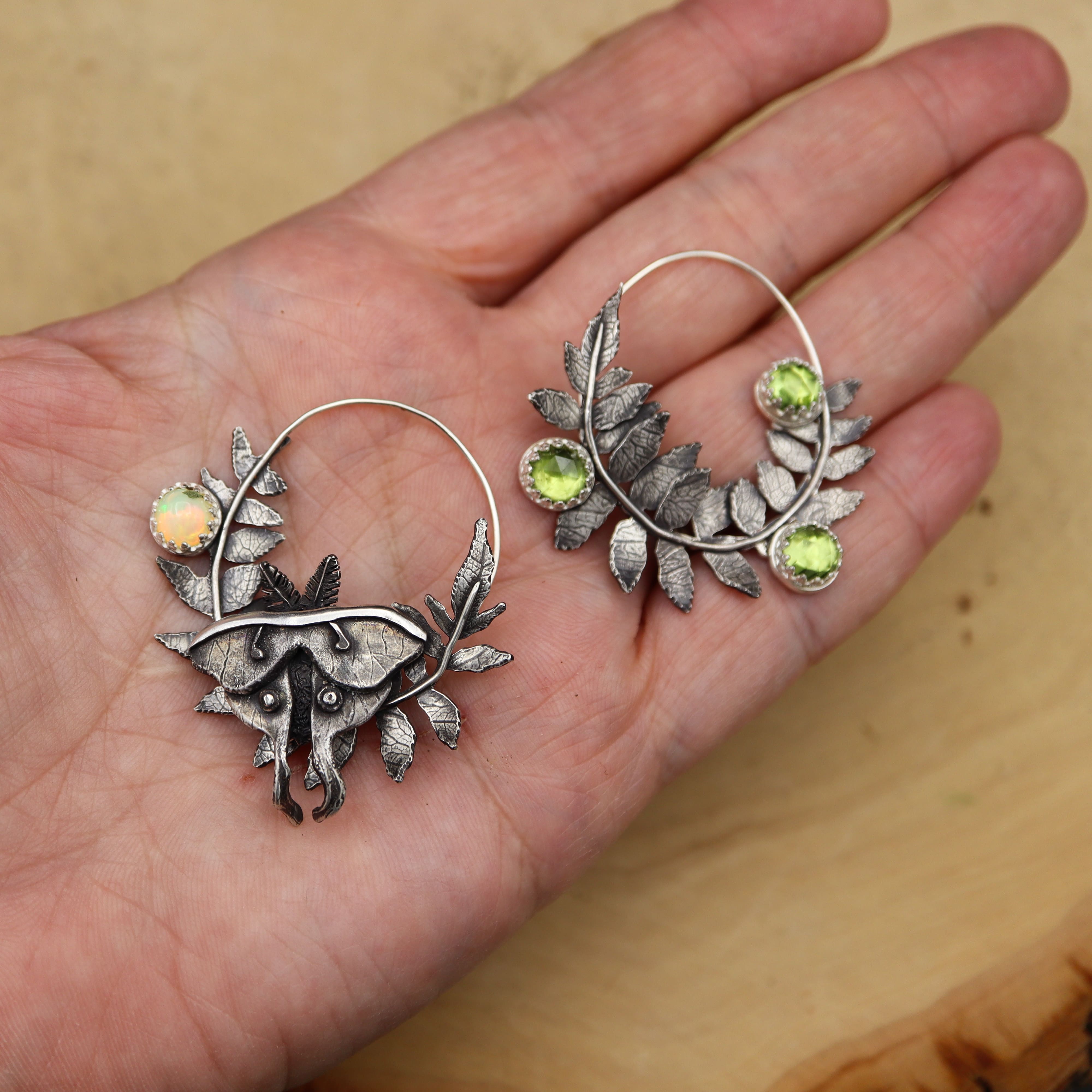 Silver staghorn sumac leaf earrings with green peridot and opal gemstones held in a hand against a wooden background