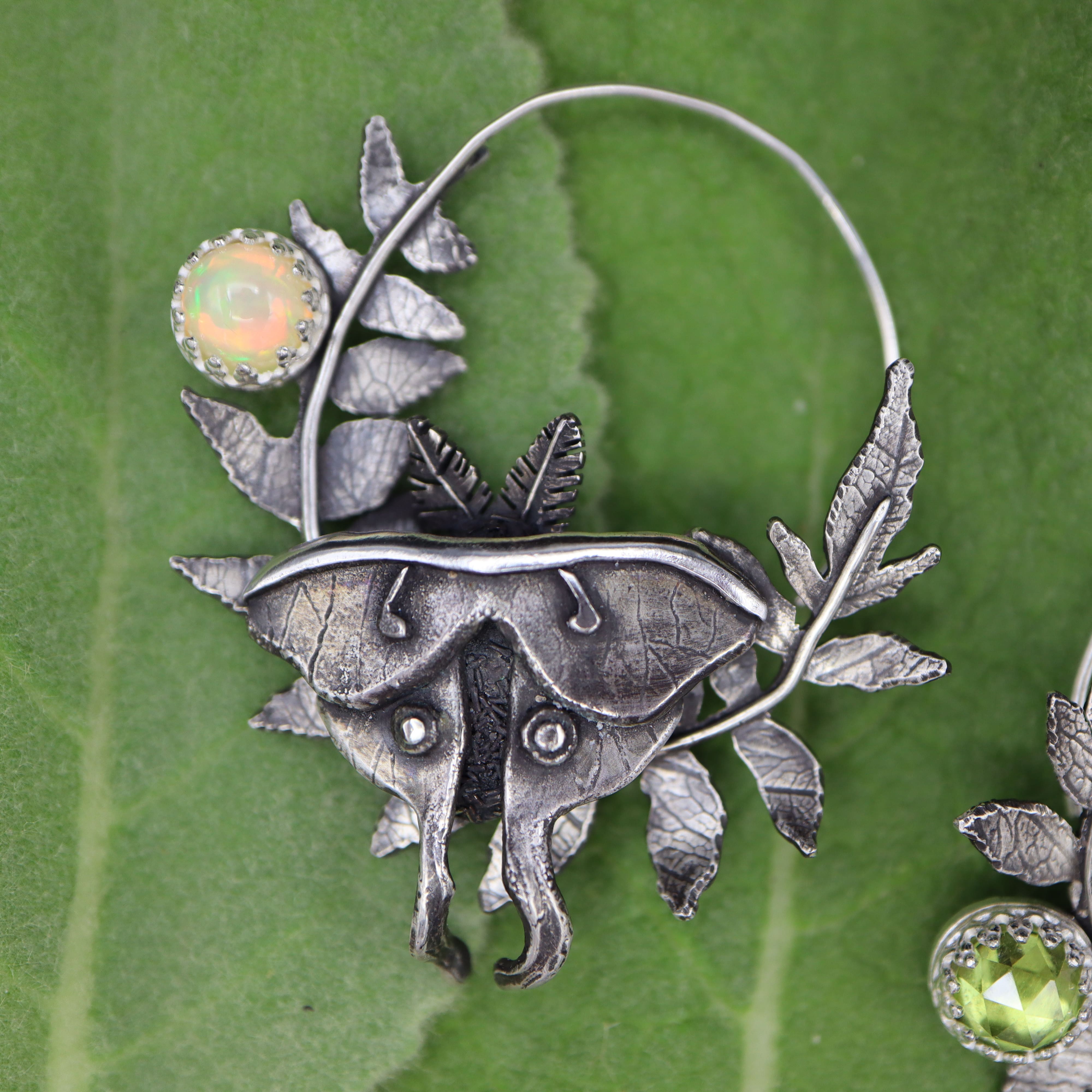 Silver luna moth earring with an opal gemstone on a green leaf background