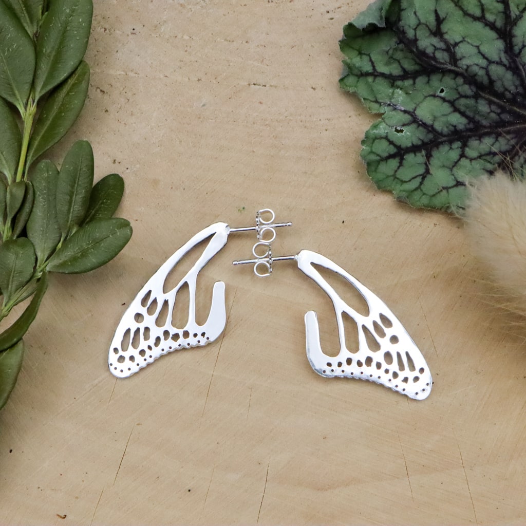 Sterling silver monarch wing earrings are shown on a piece of light tan wood and next to evergreens and wild grasses.