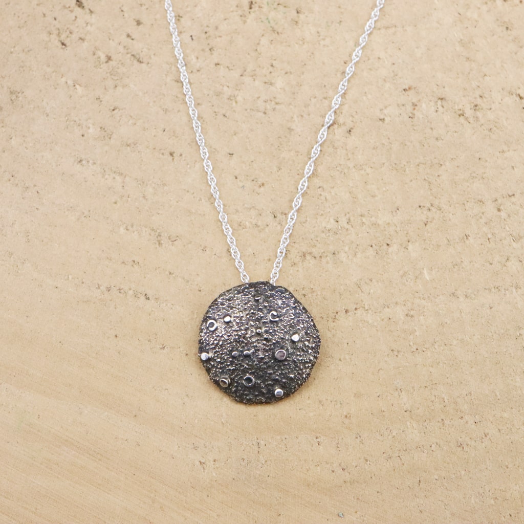 A sterling silver full moon necklace is shown on a light tan piece of wood. The moon is about 1 inch across. 
