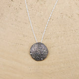 A sterling silver full moon necklace is shown on a light tan piece of wood. The moon is about 1 inch across. 