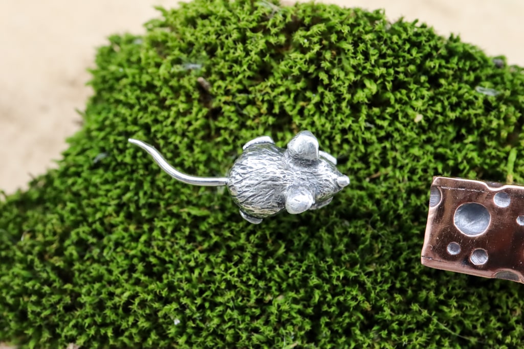 A top photo of the tiny silver mouse that was hand carved. It is shown on a piece of dark green moss. 