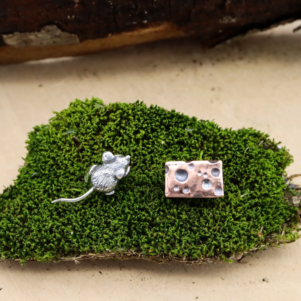 A tiny hand carved sterling silver mouse and a piece of 3 dimensional swiss cheese made from copper. They are a pair of mismatched stud earrings and shown on a piece of green moss. 