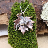 A hand carved sterling silver mouse sits in the middle of a handmade copper maple leaf. The necklace is about 1 inch tall and shown on a piece of dark green moss. 