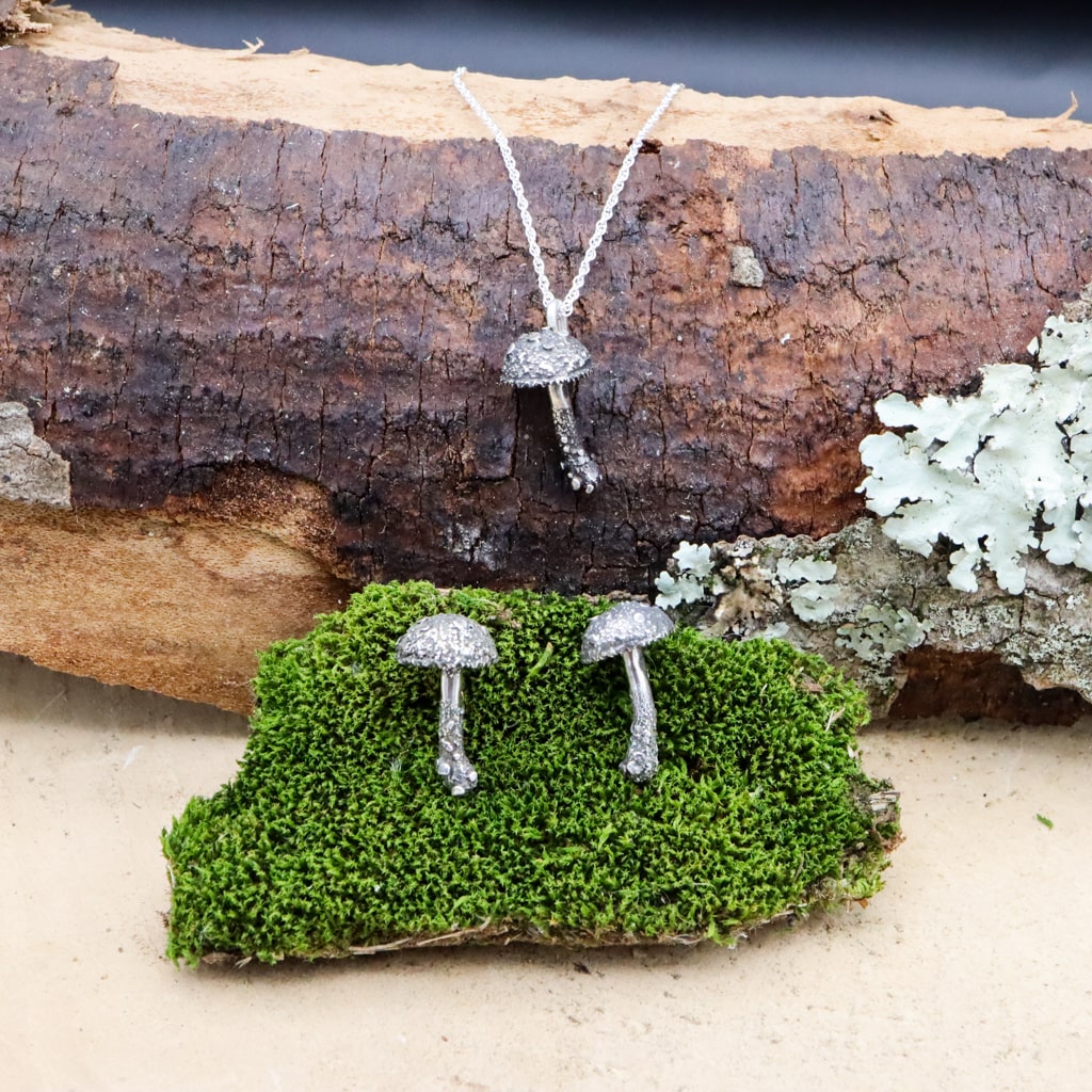 Sterling silver tiny hand carved mushroom earring studs and a matching necklace are shown on a piece of dark green moss and dark brown wood. 