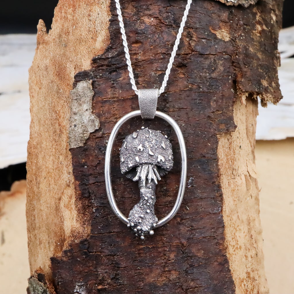 A sterling silver handmade mushroom necklace. The mushroom has a spotted cap, frilled gills, and is framed by a silver wire. The necklace is shown on a piece of tan and dark brown wood. 