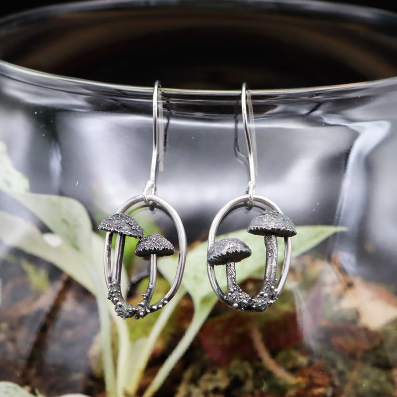 Sterling silver handmade mushroom earrings. Each earring is an oval frame with two mushrooms on each earring. They are shown hanging on a glass bowl. 