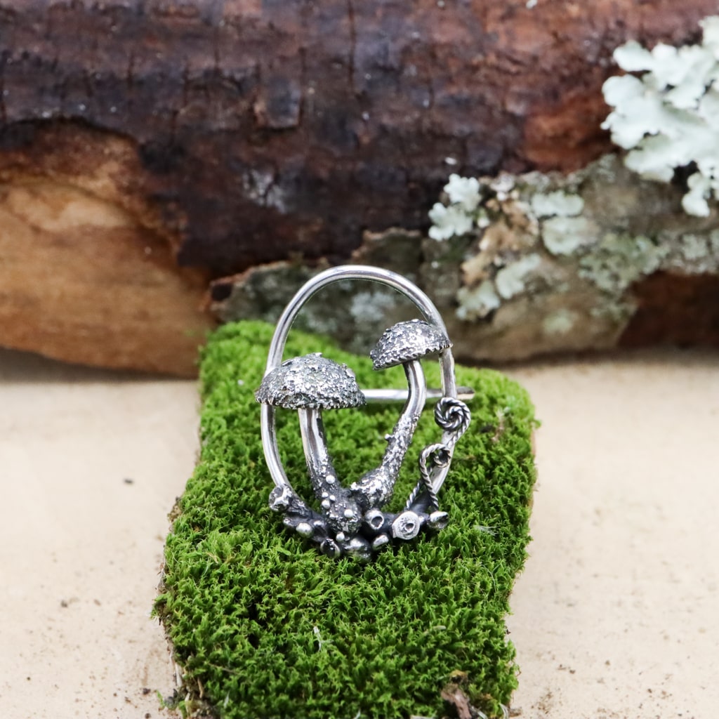 A sterling silver mushroom ring with two mushrooms inside an oval frame. There are little silver fern fronds and silver stones around the mushrooms. The ring is shown on a piece of dark green moss. 