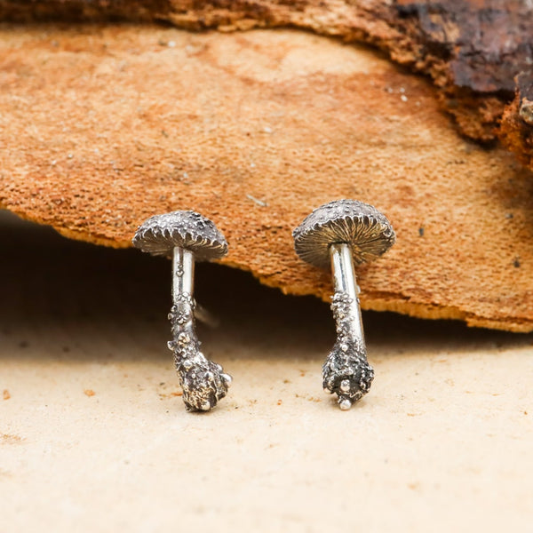 Sterling silver hand carved mushroom earrings studs. They are about 1/2 inch tall and shown on a light tan piece of bark.
