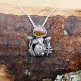 A sterling silver and watermelon tourmaline mushroom necklace. The mushroom is next to hand carved ferns, vines, and other mushrooms. The necklace is shown on a piece of dark brown wood. 