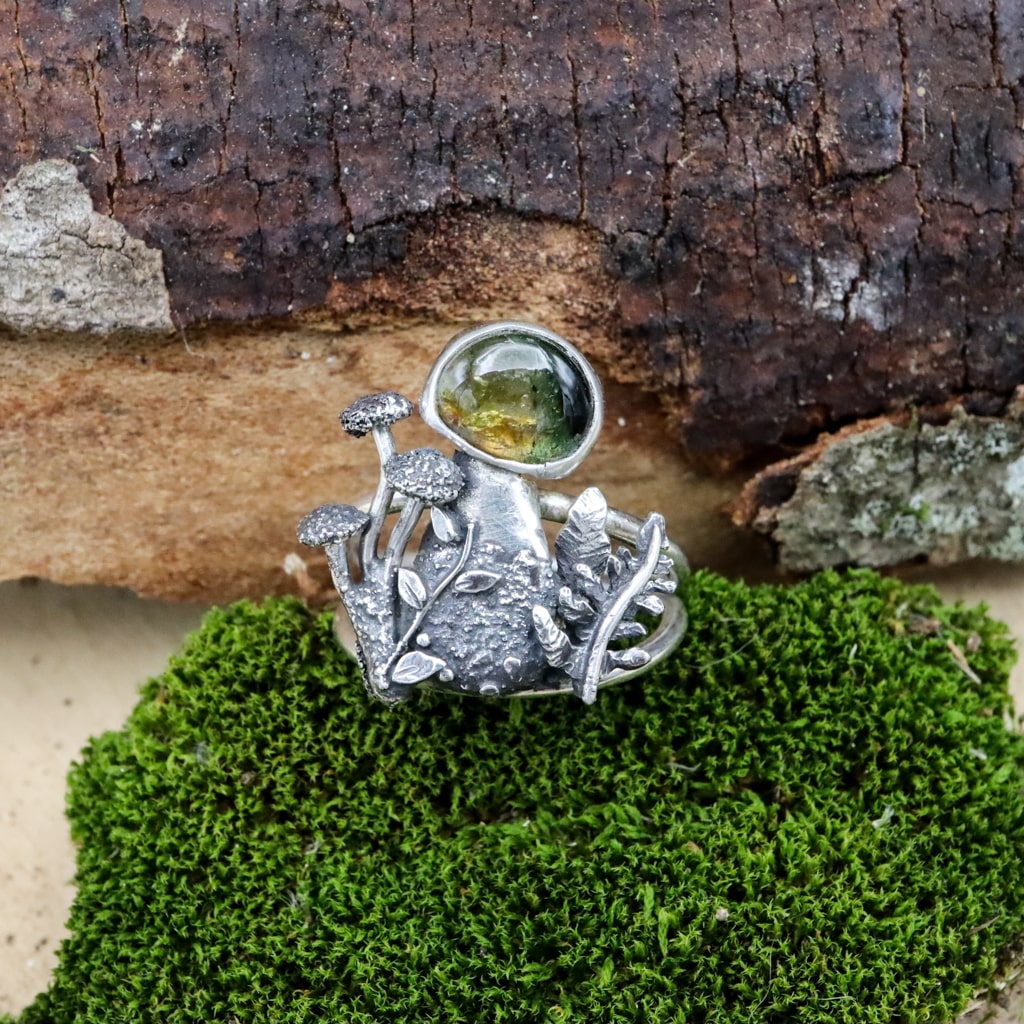 A light and dark green watermelon tourmaline gemstone sits at the top of a sterling silver mushroom ring. The mushroom is surrounded by tiny ferns, mushrooms, and leaves. It is shown on a piece of dark green moss. 