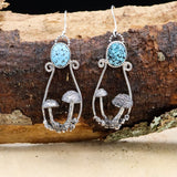 Handmade sterling silver mushroom earrings. They are shaped like hoops and each have two hand carved silver mushrooms inside the hoops. Each earrings has a light blue with black webbing turquoise stone at the top. The earrings are shown hanging on a piece of wood.