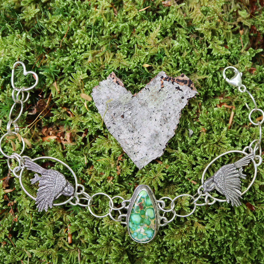 A sterling silver chickadee bracelet featuring two chickadees on each side of a spotted green Sonoran gold turquoise gemstone in the center. The bracelet is shown on some bright green moss outdoors with a heart shaped piece of bark above it. 