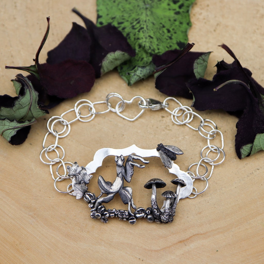 A handmade sterling silver orchid mantis bracelet. It features tiny silver mushrooms, stones, and leaves. There is a hand carved fly at the top of the bracelet. This bracelet is 7.5 inches long and shown on a light tan piece of wood with dark purple and green leaves in the background. 