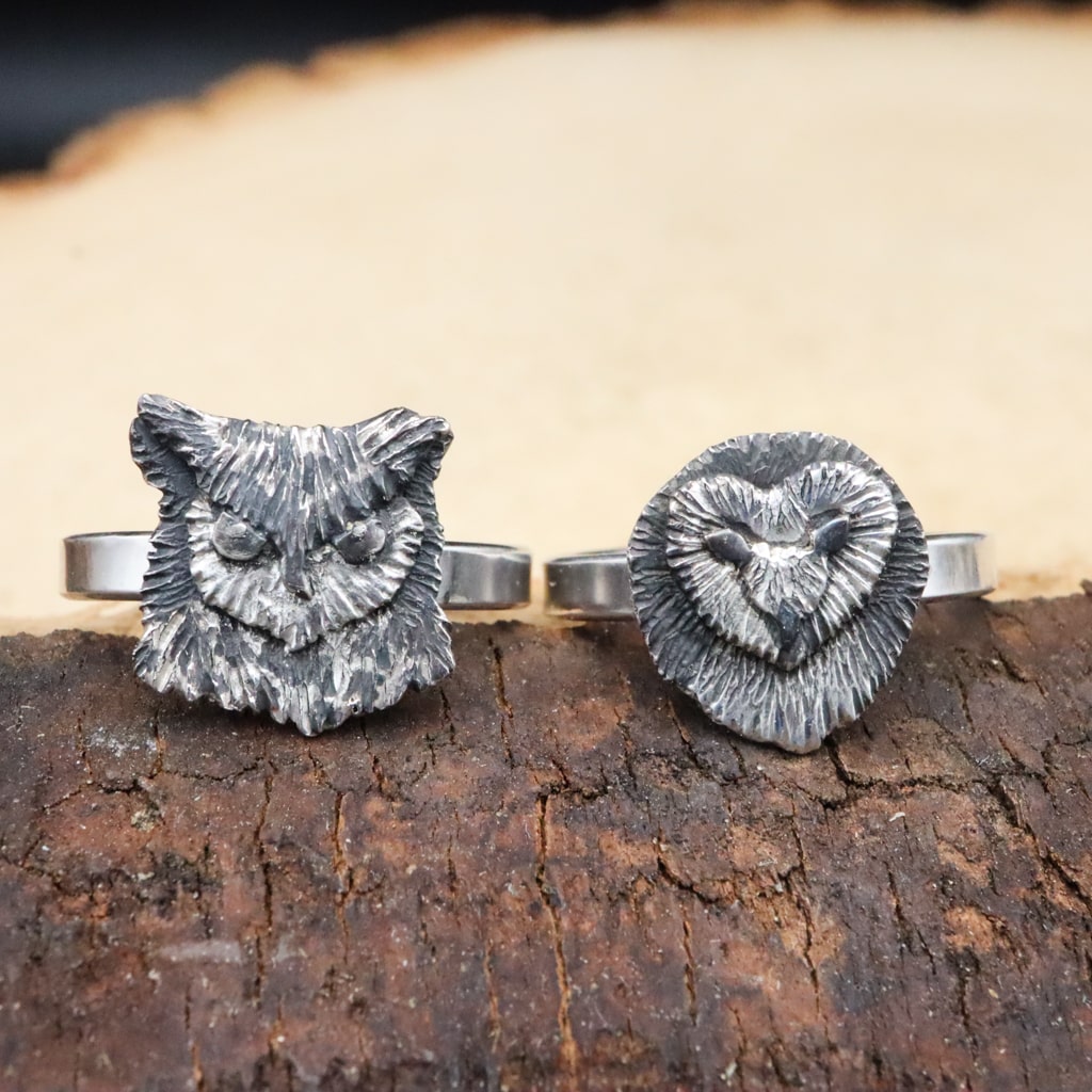 Handmade sterling silver dainty owl face ring. These silver women's rings are shown on top of a dark brown piece of wood. One ring is a great horned owl and the other ring is a barn owl.