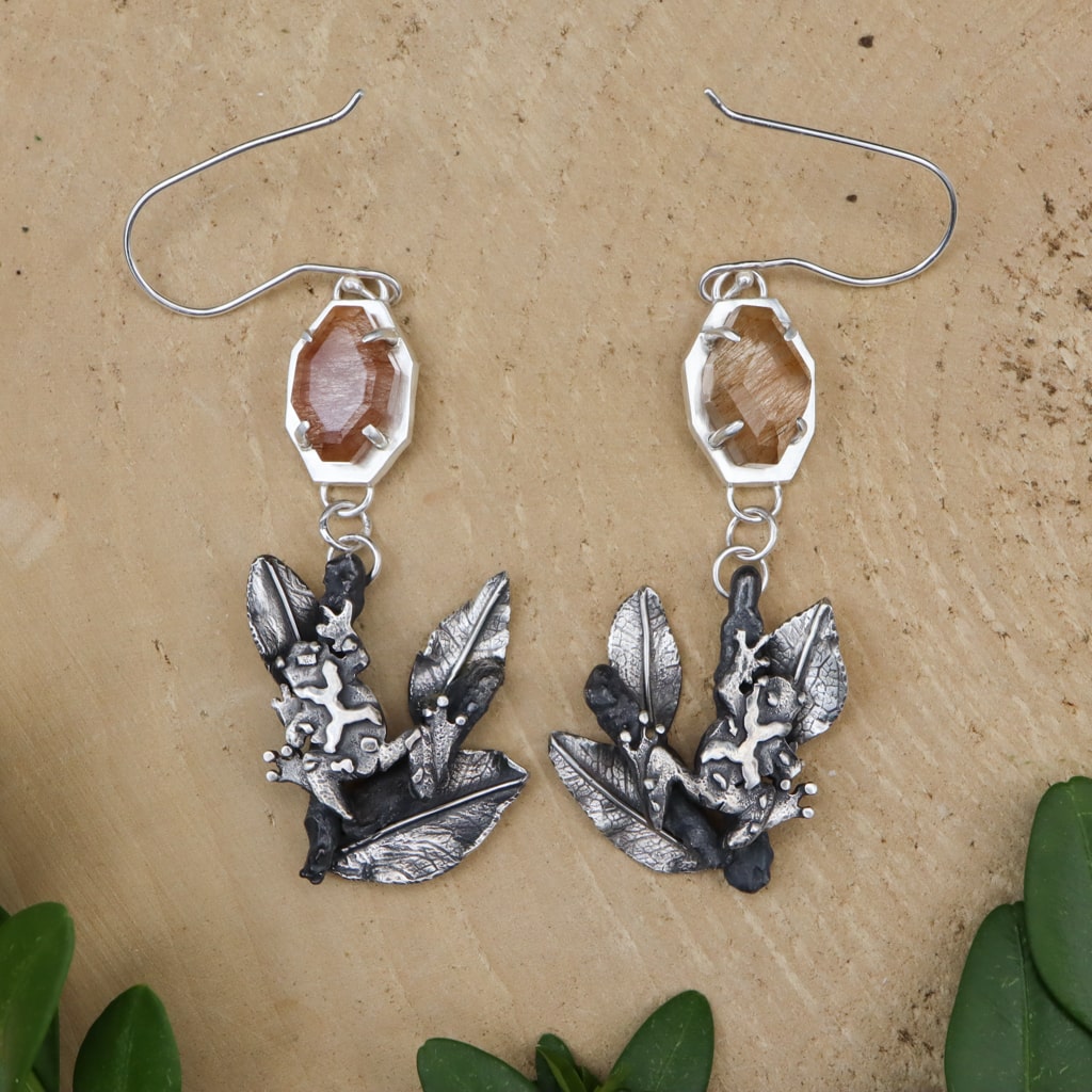 Handmade sterling silver spring peeper frog dangle earrings. There are tan colored rutilated agate stones at the top of the earrings and the frogs on branches are at the bottom. The earrings are shown on a piece of light tan wood. 