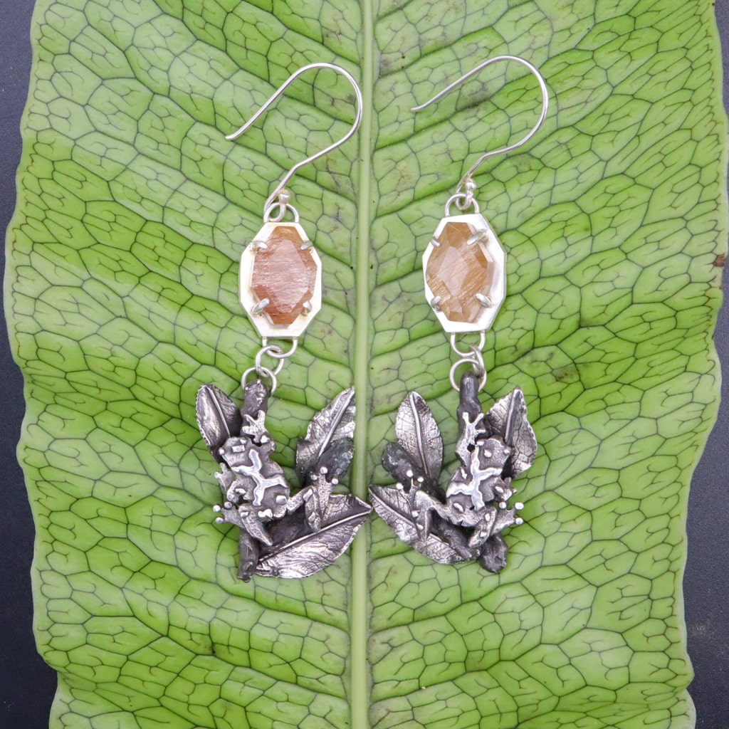 Handmade sterling silver spring peeper frog dangle earrings. There are tan colored rutilated agate stones at the top of the earrings and the frogs on branches are at the bottom. The earrings are shown on a bright green fern frond.
