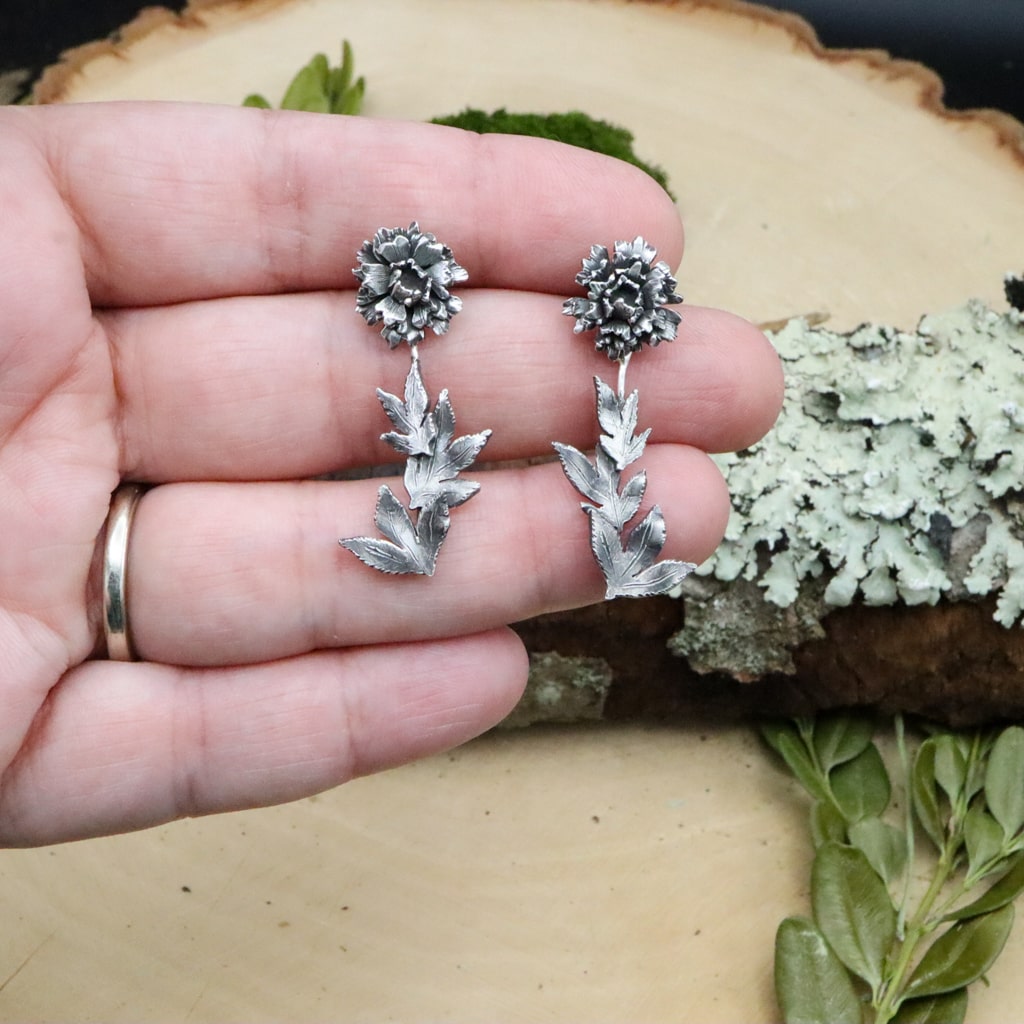A hand is shown holding the handmade sterling silver peony flower stud earrings. 