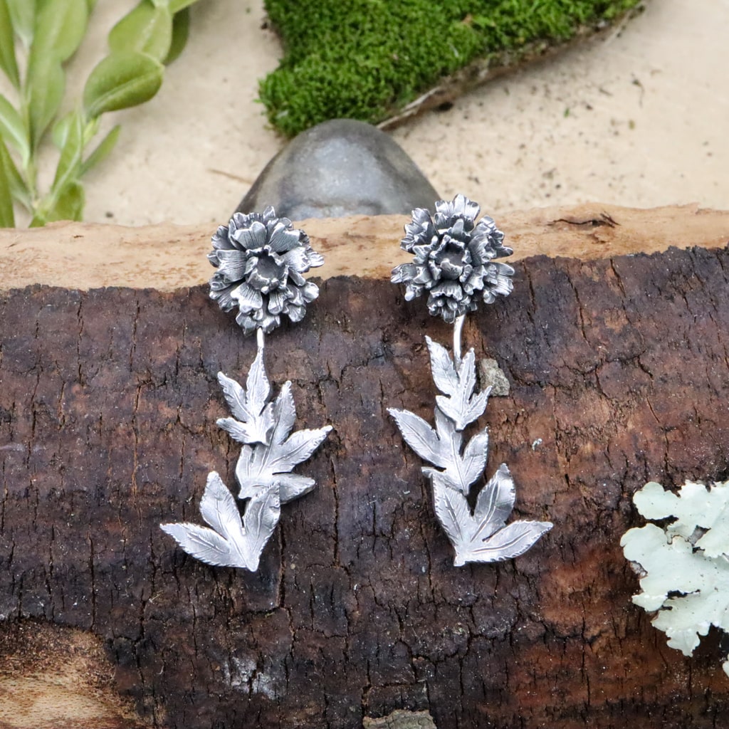 Sterling silver peony flower earrings are shown on a piece of dark brown wood. They are about 1.5 inches long and the leaf stem dangles below your earlobe. 