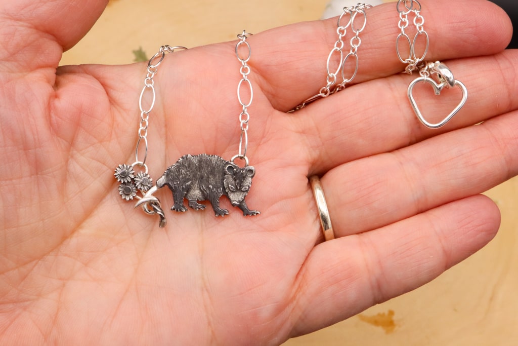 A hand is shown holding the possum necklace for size reference. It is about 1.5 inches wide and comes on an 18 inch long sterling silver necklace.